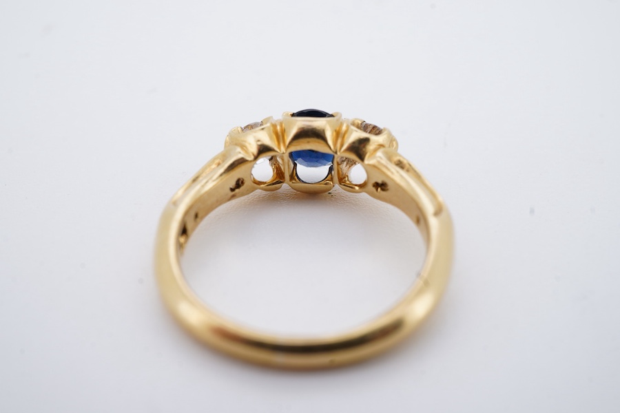 A Victorian sapphire and diamond three-stone ring, late 19th century, set with an oval sapphire between cushion-shaped diamonds totalling approximately 0.30 carats, to a gold mount of scrolled design, size J, maker's mar